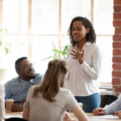 African American woman standing and talking about new project, opinion, business strategy at briefing, company meeting, multiracial colleagues group looking attentively at speaking businesswoman