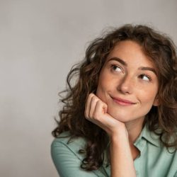 Portrait of young thoughtful woman with hand on chin having an idea against grey background. Beautiful pensive woman looking away while thinking. Close up face of natural girl planning her future isolated on gray wall with copy space.