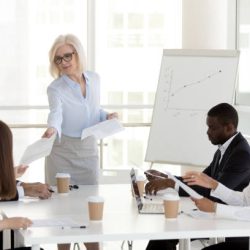 Smiling mature female manager company leader gives handout to diverse employees team at office group meeting, middle aged businesswoman handing paper finanical report to workers at corporate briefing